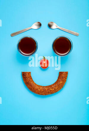 Konzept von Licht und gesunde Ernährung Frühstück. Türkische Bagel (simit) Tee Gläser, Kaffee Löffel und eine Kirsche Tomate als Smiley entworfen. Stockfoto