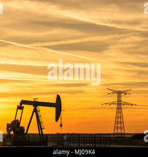 Pumpjack und Sendetürme bei Sonnenuntergang symbolisieren ökologischen Übergang. Ein Pumpenheber mit Strompylon und Stromleitung gegen einen roten Himmel. Stockfoto