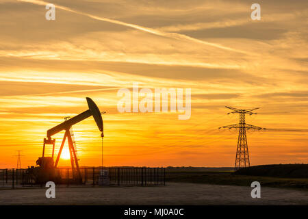 Pumpjack und Sendetürme bei Sonnenuntergang symbolisieren ökologischen Übergang. Eine Pumpenbuchse mit Strommasten und Stromleitung gegen einen roten Himmel. Stockfoto