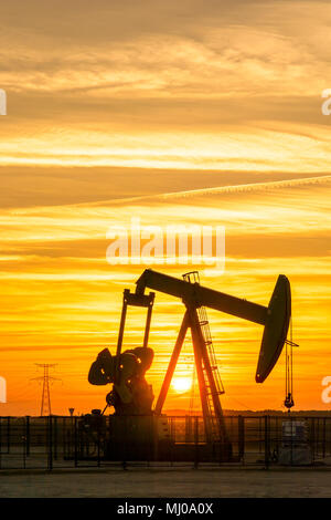 Pumpjack und Sendetürme bei Sonnenuntergang symbolisieren ökologischen Übergang. Eine Pumpenbuchse mit Strommasten und Stromleitung gegen einen roten Himmel. Stockfoto