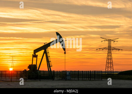 Pumpjack und Sendetürme bei Sonnenuntergang symbolisieren ökologischen Übergang. Eine Pumpenbuchse mit Strommasten und Stromleitung gegen einen roten Himmel. Stockfoto