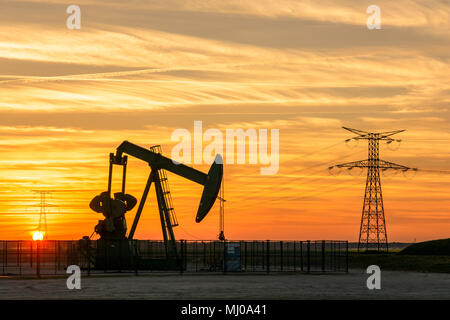 Pumpjack und Sendetürme bei Sonnenuntergang symbolisieren ökologischen Übergang. Eine Pumpenbuchse mit Strommasten und Stromleitung gegen einen roten Himmel. Stockfoto