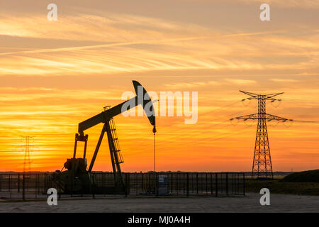 Pumpjack und Sendetürme bei Sonnenuntergang symbolisieren ökologischen Übergang. Eine Pumpenbuchse mit Strommasten und Stromleitung gegen einen roten Himmel. Stockfoto