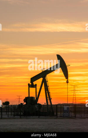 Pumpjack und Sendetürme bei Sonnenuntergang symbolisieren ökologischen Übergang. Eine Pumpenbuchse mit Strommasten und Stromleitung gegen einen roten Himmel. Stockfoto