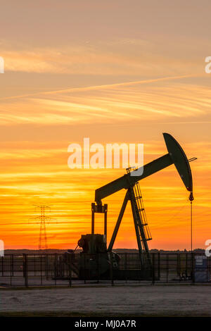 Pumpjack und Sendetürme bei Sonnenuntergang symbolisieren ökologischen Übergang. Eine Pumpenbuchse mit Strommasten und Stromleitung gegen einen roten Himmel. Stockfoto