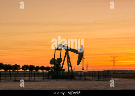Ein Kürbicker pumpt bei Sonnenuntergang Öl aus einem Brunnen Mit einer Reihe von Bäumen auf der linken Seite und ein Reihe von Sendetürmen auf der rechten Seite gegen roten Himmel Stockfoto