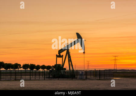 Ein Kürbicker pumpt bei Sonnenuntergang Öl aus einem Brunnen Mit einer Reihe von Bäumen auf der linken Seite und ein Reihe von Sendetürmen auf der rechten Seite gegen roten Himmel Stockfoto