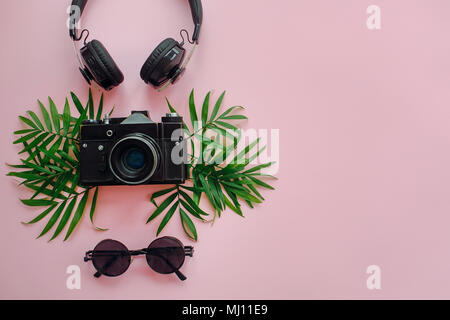 Elegante schwarze Foto Kamera mit grünen Palmenblättern, Sonnenbrille und Kopfhörer am trendigen rosa Hintergrund, flach. Moderne hipster Reisen und Fernweh Stockfoto