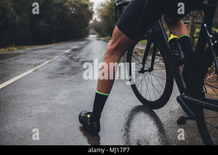 Niedrige Abschnitt Schuß von männlichen Athleten, der mit seinem Fahrrad. Man Radfahrer mit seinem Fahrrad auf nasser Straße. 7/8 shot der männlichen Triathlet mit seinem cy Stockfoto