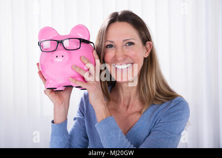 Lächelnd reife Frau mit rosa Sparschwein mit schwarzen Brillen gegen weißen Jalousien Stockfoto
