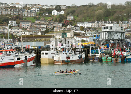 Die traditionelle Cornish racing gig Pendeen Ausbildung in Newlyn Harbour in den Vordergrund mit Fischereifahrzeugen und die Stadt von Newlyn im Hintergrund. Stockfoto
