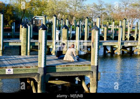 Ein junger Mann und Frau in ihren zwanziger Jahren sitzen auf einem Boot Dock mit Blick auf der Chesapeake Bay im Downtown Annapolis Harbour, MD, USA Stockfoto