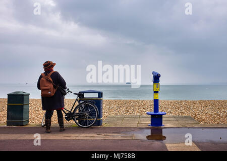 Frau mit Blick auf das Meer mit dem Fahrrad neben sprechen Teleskop auf der Promenade in Fareham, Hampshire an einem bewölkten Tag Stockfoto