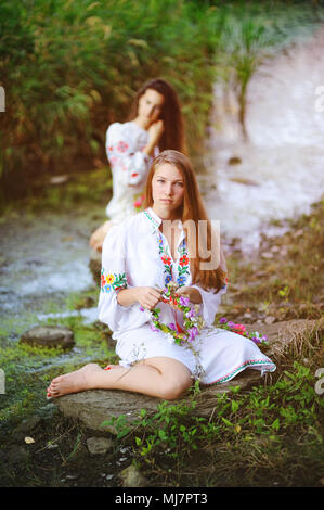 Zwei junge schöne Mädchen in weißem Hemd mit floralen Ornament mit Kränze in ihren Händen sitzen auf dem Hintergrund des Flusses. Stockfoto