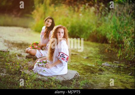 Zwei junge schöne Mädchen in weißem Hemd mit floralen Ornament mit Kränze in ihren Händen sitzen auf dem Hintergrund des Flusses. Stockfoto