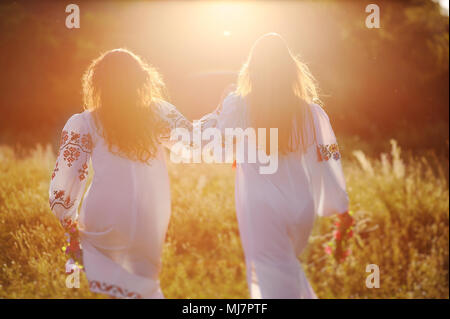 Zwei junge schöne Mädchen in weißen Hemden mit floralen Ornament mit Blumenkranz in den Händen laufen vor dem Hintergrund der Natur und Gras in der Kontur oder im Gegenlicht der Sonne. Stockfoto