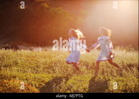 Zwei junge schöne Mädchen in weißen Hemden mit floralen Ornament mit Blumenkranz in den Händen laufen vor dem Hintergrund der Natur und Gras in der Kontur oder im Gegenlicht der Sonne. Stockfoto