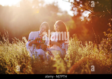 Zwei jungen hübschen Mädchen in weißen Hemden Geflecht sich gegenseitig und Webart Kränze von Blumen vor dem Hintergrund des Grases und Natur bei Sonnenuntergang. Stockfoto