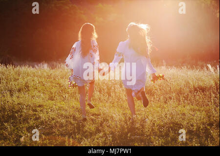 Zwei junge schöne Mädchen in weißen Hemden mit floralen Ornament mit Blumenkranz in den Händen laufen vor dem Hintergrund der Natur und Gras in der Kontur oder im Gegenlicht der Sonne. Stockfoto