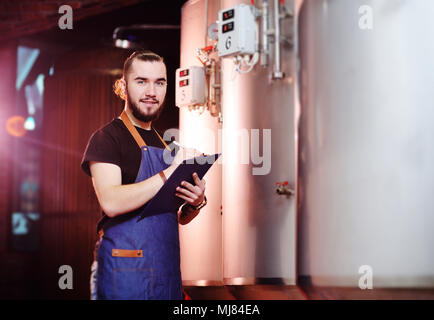 Bärtige Brauer Mann auf einem Hintergrund von Lagertanks und eine Brauerei Notizen in einem Tablet Stockfoto