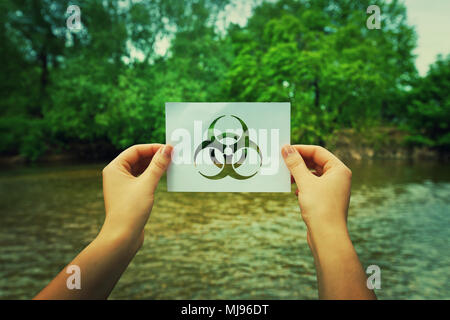 Frau Hände halten ein Blatt Papier mit einer Infektion Symbol in der Mitte der Natur in der Nähe von einem Fluss und den Wald. Biohazard Zeichen, Warnung Virus Stockfoto