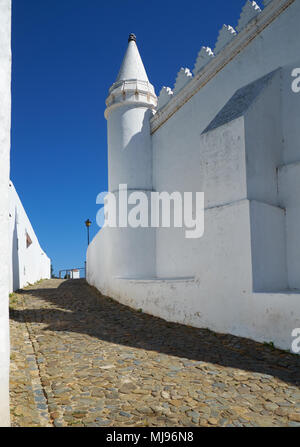 Der Blick auf die Straße mit Pflastersteinen und die weiße Wand der Kirche (Igreja Matriz) entlang in Mertola. Baixo Alentejo. Portugal Stockfoto