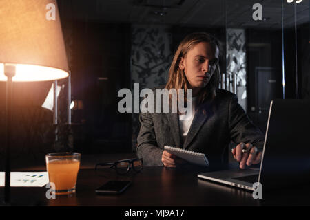 Müde Geschäftsmann durch den zeitgenössischen Laptop in der Nacht die Arbeit im Büro. Hipster mit langen Haaren halten Notizblock in der Hand und nutzt mobile Computer. Film Effek Stockfoto