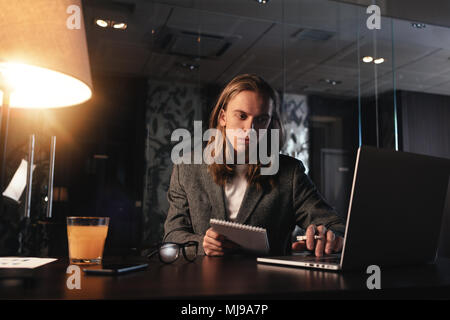 Müde junge Mann mit langen Haaren in der Nacht loft Büro. Mitarbeiter arbeiten in der modernen coworking Studio. Geschäftsmann nutzt moderne Laptop Stockfoto