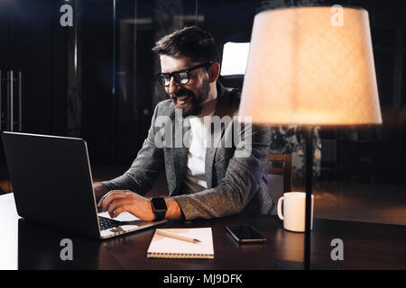 Lächelnde junge Bartgeier Geschäftsmanns, der auf zeitgenössische Notebook im Loft Office in der Nacht. Projekt Manager Brille Text eingeben auf Laptop Stockfoto