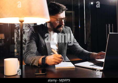 Bartgeier Projekt Manager halten Sie Stift in der Hand und mit zeitgenössischen Notebook. Junge Unternehmer im Loft Office in der Nacht arbeiten. Mann, der neue Anlauf Stockfoto