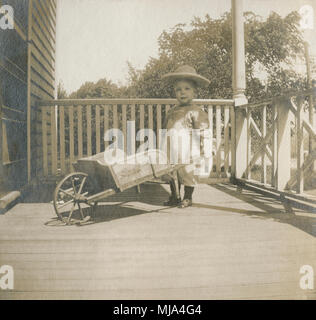 Antike c 1902 Foto, Kleinkind Junge in viktorianischer Kleidung mit Schubkarre auf der Veranda. Lage unbekannt, wahrscheinlich New England, USA. Quelle: ORIGINAL FOTOABZUG. Stockfoto