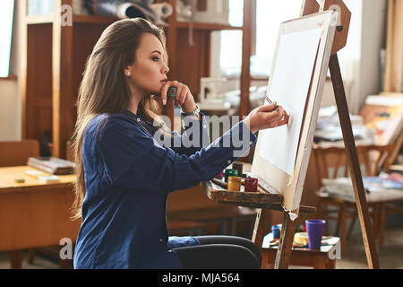 Junge schöne Mädchen, weibliche Künstler Maler denken an ein neues Artwork Idee und bereit ist, den ersten Pinselstrich auf Holz Staffelei mit einem Pinsel in s zu machen Stockfoto