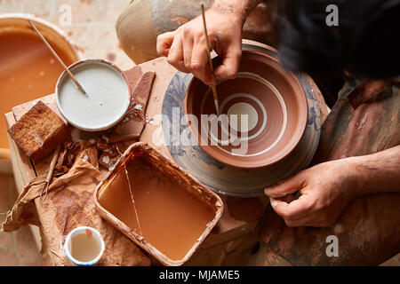Ein Potter in einem Plaid Shirt und Schürze malt ein Ton Platte in Weiß in Werkstatt, Ansicht von oben, Nahaufnahme, selektiven Fokus. Es gibt einige Tools, Ton Proben Stockfoto