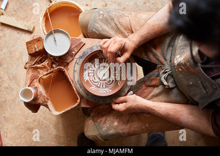 Ein Potter in einem Plaid Shirt und Schürze malt ein Ton Platte in Weiß in Werkstatt, Ansicht von oben, Nahaufnahme, selektiven Fokus. Es gibt einige Tools, Ton Proben Stockfoto