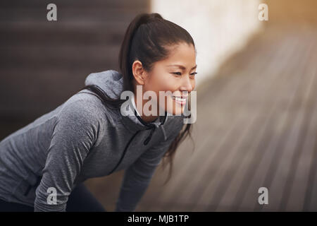 Lächelnde junge asiatische Frau stehen im freien Ausübung Kleidung tragen, mit den Händen auf den Knien die Vorbereitung für einen Lauf zu gehen Stockfoto