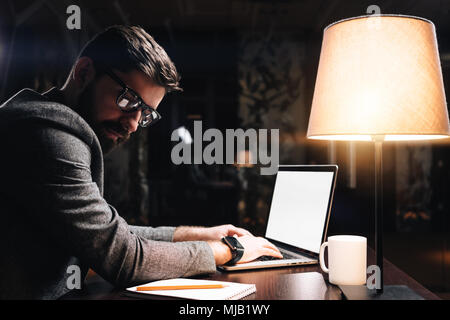 Junge Mitarbeiter Brille Text eingeben auf zeitgenössische Laptop in der modernen Loft Büro bei Nacht Bärtigen. Business Mann sitzt durch den hölzernen Tisch mit l Stockfoto
