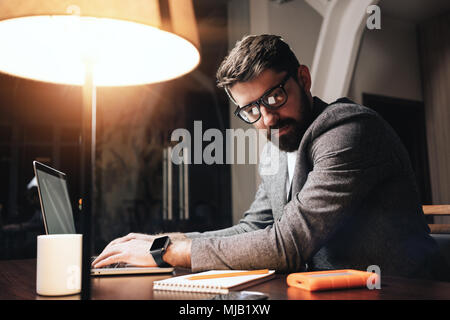 Bärtige Geschäftsmann in Brillen durch den zeitgenössischen Laptop in der Nacht Mitarbeiter Studio arbeiten. Junge Projektmanager mit der Texteingabe auf dem Notebook. Film ef Stockfoto