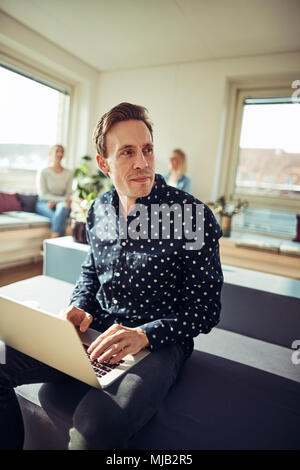 Junge Unternehmer saß auf seinem Schreibtisch, die an einem Notebook arbeitet mit Kollegen zusammen, die im Hintergrund sitzen Stockfoto