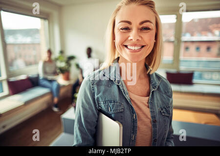 Lächelnden jungen Geschäftsfrau Holding einen Laptop unter den Arm, während in einem modernen Büro mit Kollegen sprechen im Hintergrund Stockfoto