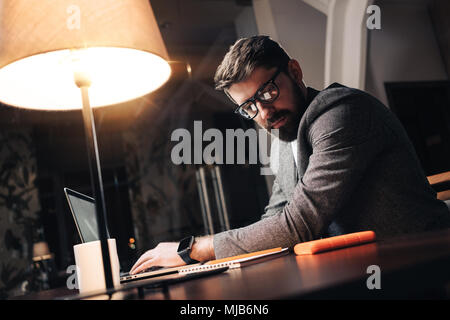 Bärtige junge Unternehmer in Gläsern sitzt durch den hölzernen Tisch mit Lampe und mit zeitgenössischen mobile Notebook im Loft Office in der Nacht. Mitarbeiter worki Stockfoto