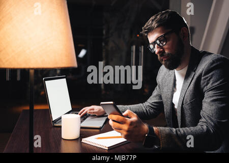 Geschäftsmann mit Smartphone und Laptop in Coworking Studio. Junger bärtiger Mann über neue Startup in der Nacht arbeiten in modernen Loft Büro. Film Effekt Stockfoto