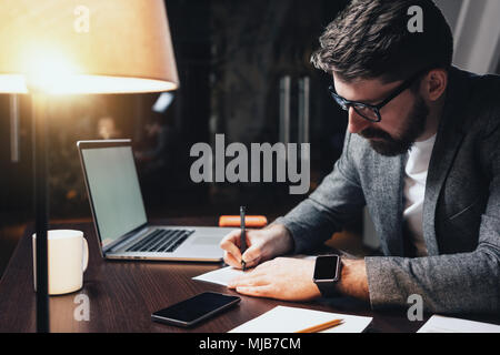 Junge bärtige Mann mit Brille, erobert und Büro mit modernen Laptop in der Nacht loft Büro. Brainstorming Prozess. Geschäftsmanns, der wit Stockfoto