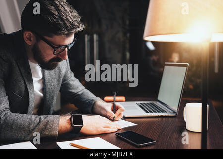 Bärtige Geschäftsmann sitzt in der Nacht loft Büro und Arbeiten mit Dokumenten und modernen Laptop. Stilvolle Mann mit Brille sitzt durch den hölzernen Tisch Stockfoto