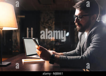 Bärtige Geschäftsmann tragen Brillen und mit pro Computer im modernen Büro in der Nacht. Mitarbeiter halten Smartphone. Horizontale. Film Effekt Stockfoto