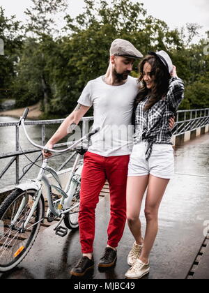 Schöne junge Paare von Mann und Frau im Sommer Kleidung gehen mit dem Fahrrad umarmen am Park in der Brücke Stockfoto