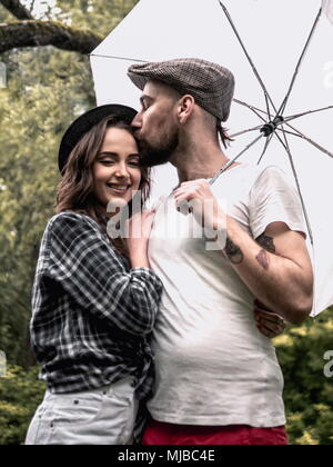 Schöne junge Paar Mann und Frau im Sommer Kleidung springen, umarmen, küssen und Lachen im Park mit wtite Regenschirm unter dem Regen Stockfoto