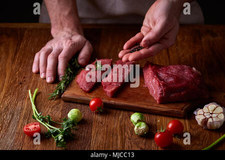 Vorbereitung für das Abendessen. Kochen, Verarbeitung von Fleisch. Die starke professionelle man Hände Verschütten von Salz auf rohen Beefsteak, selektiver Fokus, Nahaufnahme. Self-c Stockfoto