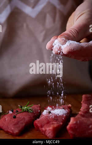 Vorbereitung für das Abendessen. Kochen, Verarbeitung von Fleisch. Die starke professionelle man Hände Verschütten von Salz auf rohen Beefsteak, selektiver Fokus, Nahaufnahme. Self-c Stockfoto