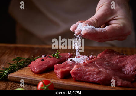 Vorbereitung für das Abendessen. Kochen, Verarbeitung von Fleisch. Die starke professionelle man Hände Verschütten von Salz auf rohen Beefsteak, selektiver Fokus, Nahaufnahme. Self-c Stockfoto