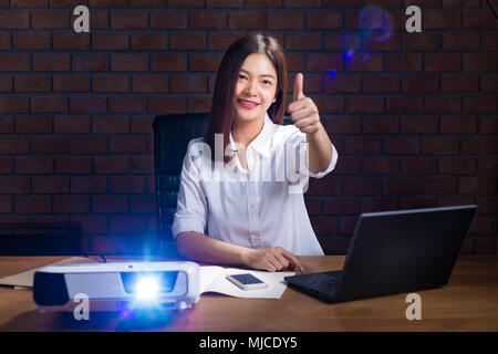 Jungen asiatischen Geschäftsfrau im weißen Hemd arbeiten spät in ihrem Büro beobachten business Präsentation durch Ihren Projektor und glücklich einen Daumen nach oben Stockfoto